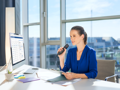 Woman in a blue shirt sitting at a desk, dictating with Philips SpeechMike into a computer, in an office setting.