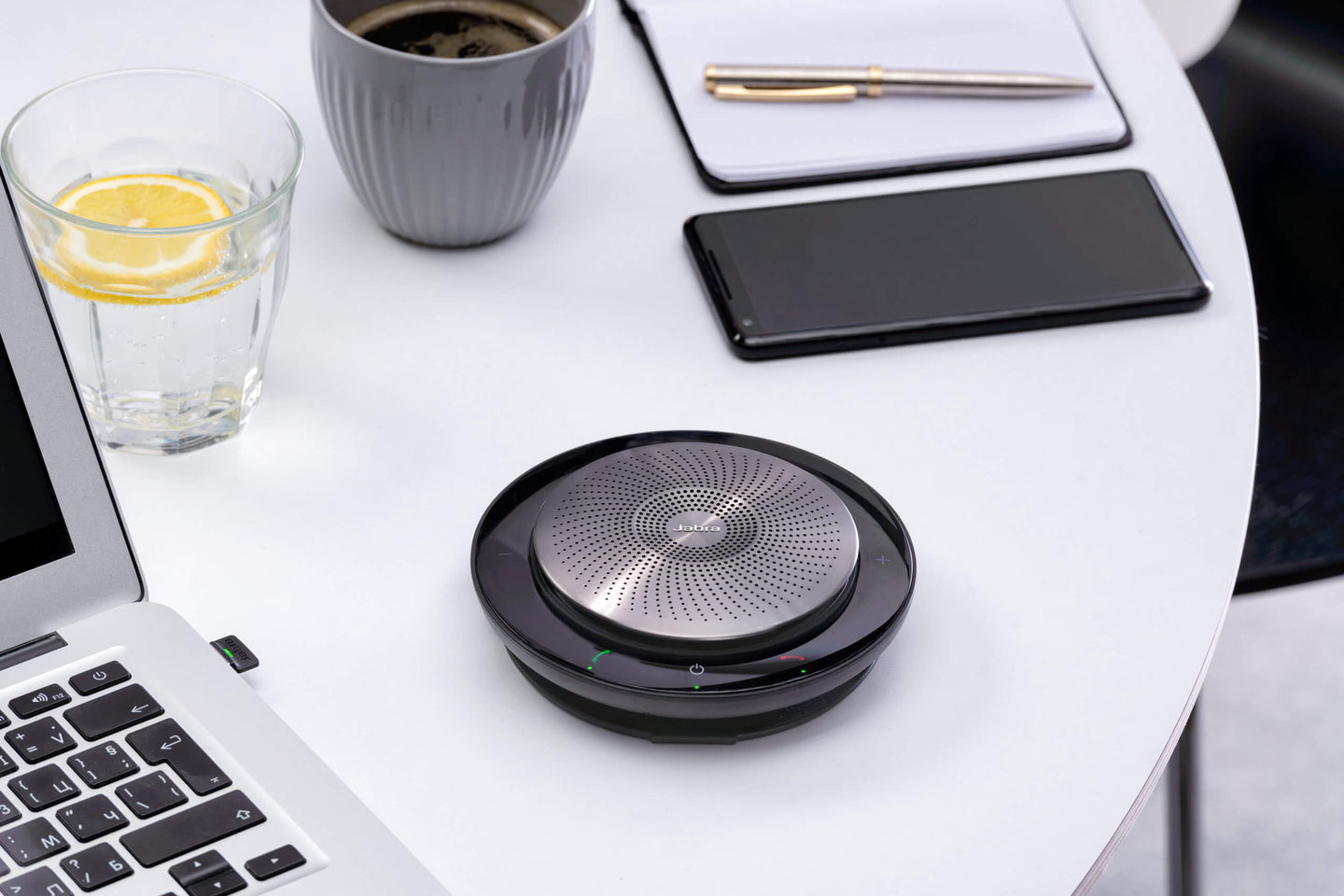 Jabra Speak 750 speakerphone on desk with laptop, smart phone, glass of water and notebook with pen