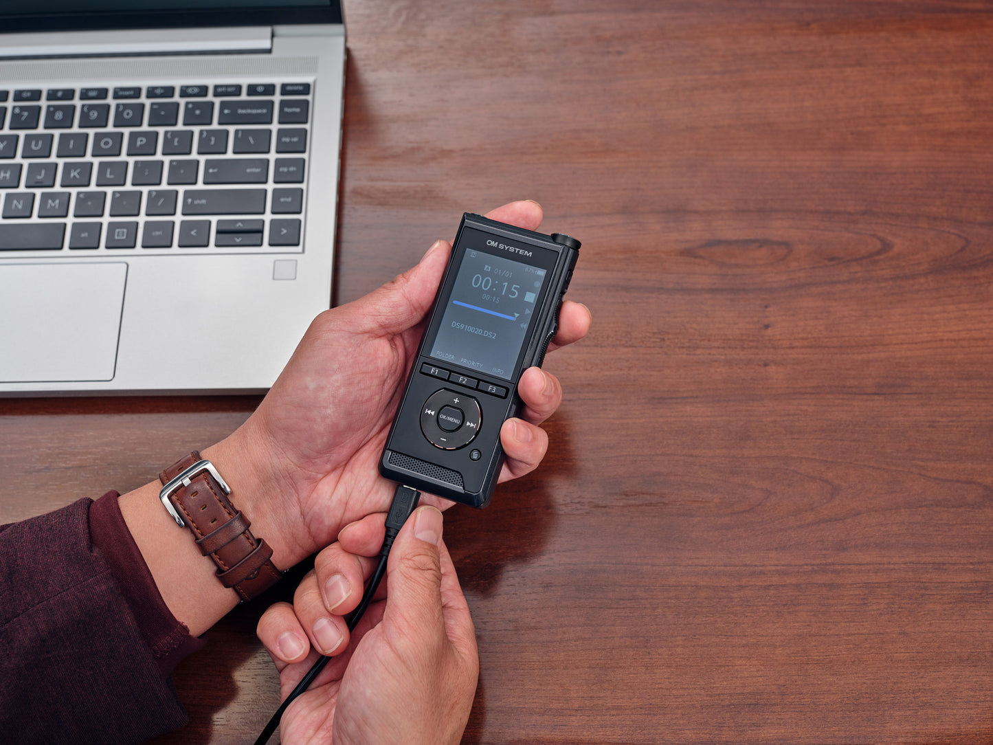 One hand holding OM System DS-9100 and second hand inserting usb cable to bottom of device to connect to computer for download